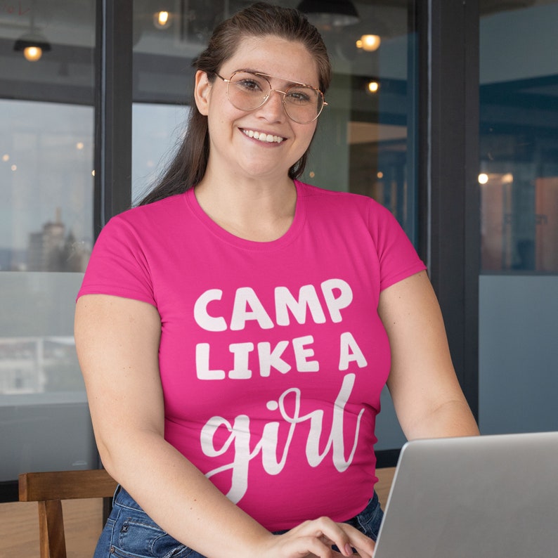 K&ouml;nnte beinhalten: Eine Frau tr&auml;gt ein pinkfarbenes T-Shirt mit dem Text "CAMP LIKE A girl" in wei&szlig;en Buchstaben. Sie l&auml;chelt und schaut in die Kamera. Sie sitzt an einem Tisch mit einem Laptop vor sich.
