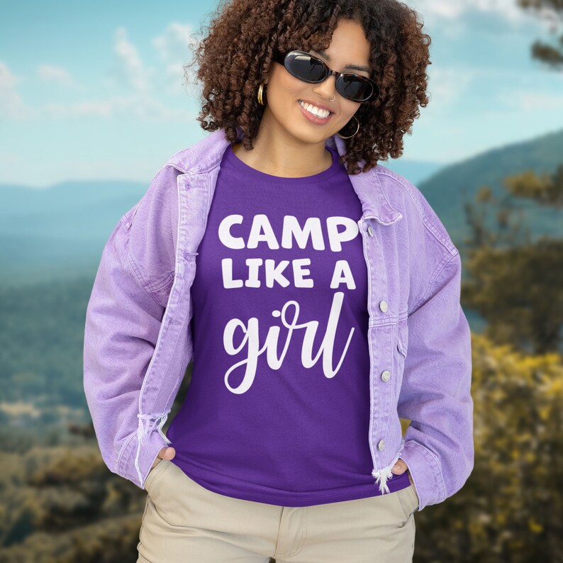 K&ouml;nnte beinhalten: Eine Frau tr&auml;gt ein lila T-Shirt mit dem Text "Camp Like A Girl" in wei&szlig;en Buchstaben. Sie tr&auml;gt eine helllila Jeansjacke und eine Khakihose. Sie steht vor einem Bergpanorama.