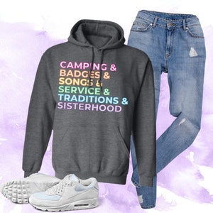 以下が含まれることがあります： レインボーカラーで「Camping & Badges & Songs & Service & Traditions & Sisterhood」と書かれたグレーのフード付きスウェットシャツです。