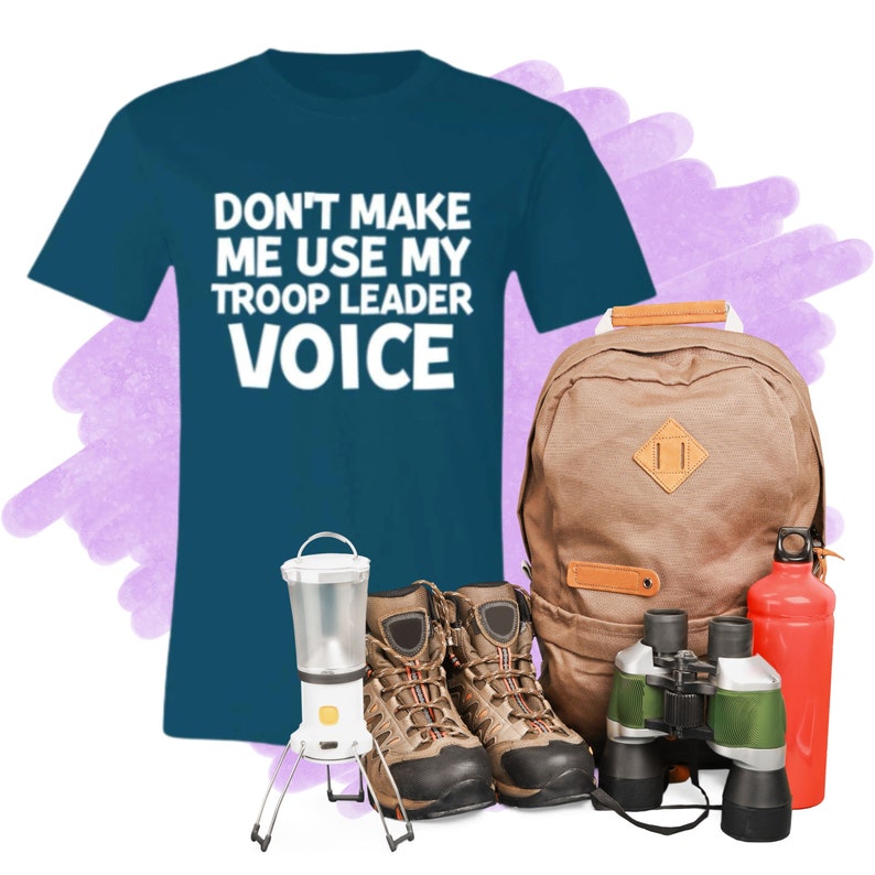 May include: A teal blue t-shirt with the text "Don't make me use my troop leader voice" in white. The shirt is next to a brown backpack, a pair of brown hiking boots, a lantern, binoculars, and a red water bottle.