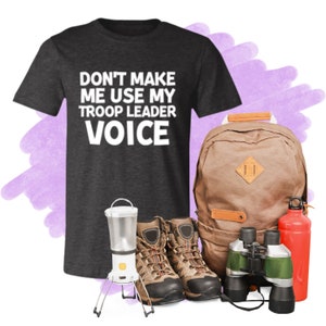 May include: A dark grey t-shirt with the text "Don't make me use my troop leader voice" in white. The shirt is next to a brown backpack, a pair of brown hiking boots, a green and silver pair of binoculars, and a red water bottle.