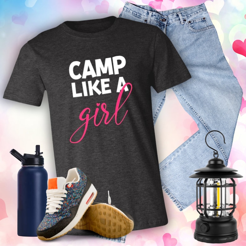 Puede incluir: Una camiseta gris oscuro con el texto "CAMP LIKE A girl" en letras blancas y rosas. La camiseta se combina con unos vaqueros azules, una botella de agua y una linterna negra.
