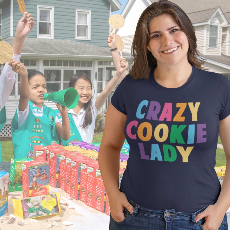 May include: A woman wearing a navy blue t-shirt with the text "CRAZY COOKIE LADY" in rainbow colours. She is standing in front of a table with Girl Guide cookies and Girl Guides selling cookies.