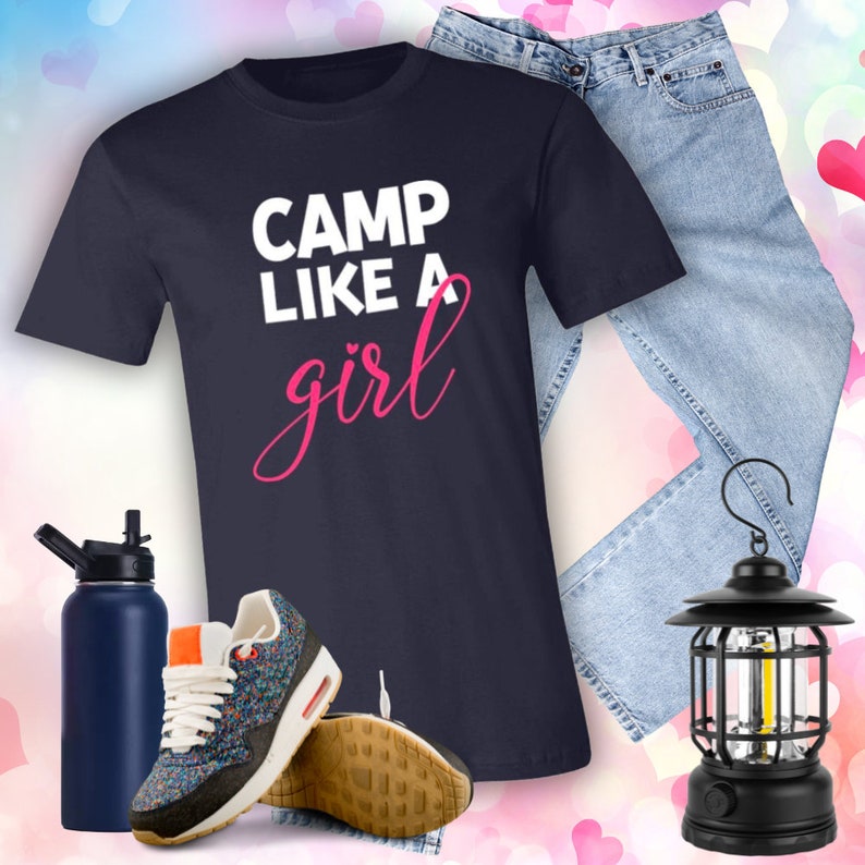 Puede incluir: Una camiseta azul marino con el texto "Camp Like A Girl" en letras blancas y rosas. La camiseta se combina con unos vaqueros azules, una botella de agua, zapatillas deportivas y una linterna negra.