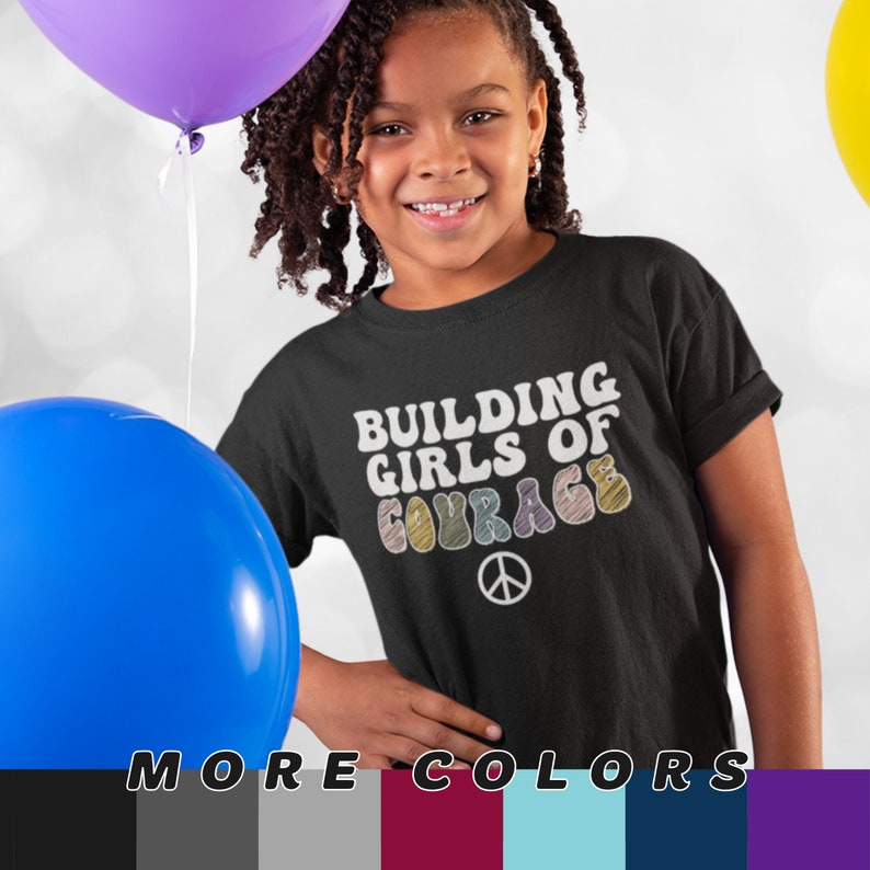 May include: A black t-shirt with the text "BUILDING GIRLS OF COURAGE" in white and colourful letters. A peace symbol is below the text. The shirt is worn by a smiling child, with balloons in the background.