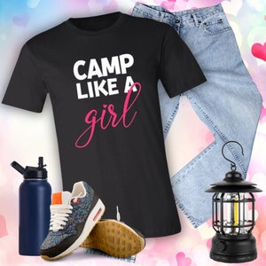 Puede incluir: Camiseta negra con el texto "Camp Like A Girl" en letras blancas y rosas. La camiseta se combina con unos vaqueros azules, una botella de agua y una linterna negra.