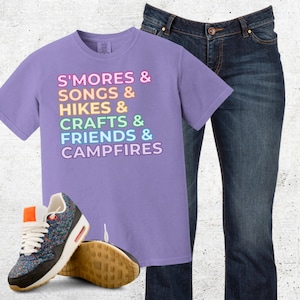 Puede incluir: Una camiseta lila con las palabras "S'MORES & SONGS & HIKES & CRAFTS & FRIENDS & CAMPFIRES" en texto colorido. Se combina con vaqueros oscuros y zapatillas deportivas estampadas con suela marr&oacute;n.