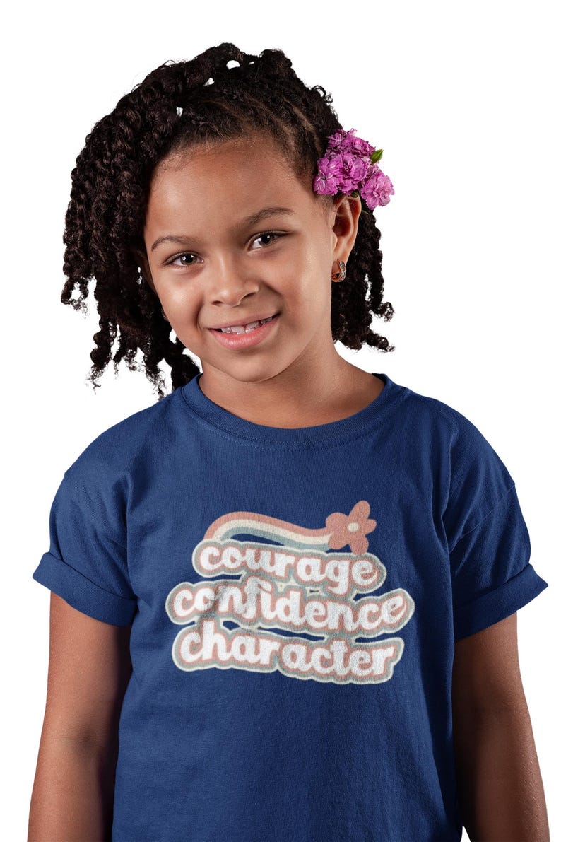 Puede incluir: Una persona joven lleva una camiseta azul marino con un gr&aacute;fico de estilo retro que dice "coraje confianza car&aacute;cter" en un esquema de colores arco&iacute;ris.