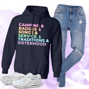 以下が含まれることがあります： レインボーカラーのグラフィックが施されたネイビーブルーのパーカー。パーカーには「Camping & Badges & Songs & Service & Traditions & Sisterhood」の文字が書かれています。パーカーはライトブルーのジーンズと白いスニーカーと合わせています。