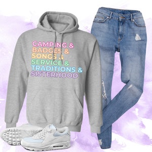 以下が含まれることがあります： レインボーカラーで「Camping & Badges & Songs & Service & Traditions & Sisterhood」とプリントされたグレーのフード付きスウェットシャツです。