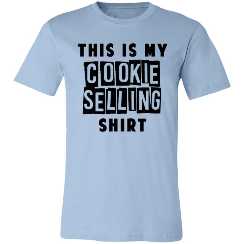 May include: Light blue t-shirt with black text that reads "This is my cookie selling shirt."