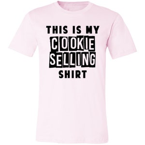 May include: Pink t-shirt with black text that reads "This is my cookie selling shirt."