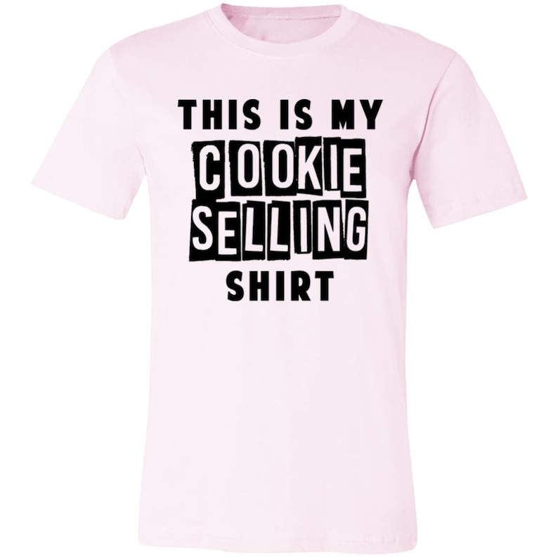 May include: Pink t-shirt with black text that reads "This is my cookie selling shirt."