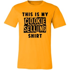May include: A yellow t-shirt with black text that reads "This is my cookie selling shirt".