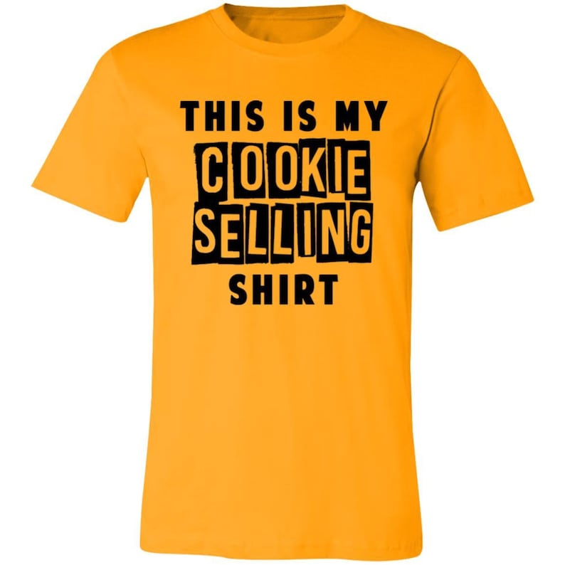 May include: A yellow t-shirt with black text that reads "This is my cookie selling shirt".