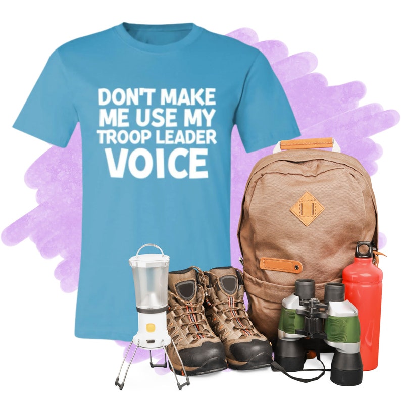 May include: A light blue t-shirt with the text "Don't make me use my troop leader voice" in white. The shirt is next to a brown backpack, a pair of brown hiking boots, a lantern, binoculars, and a red water bottle.