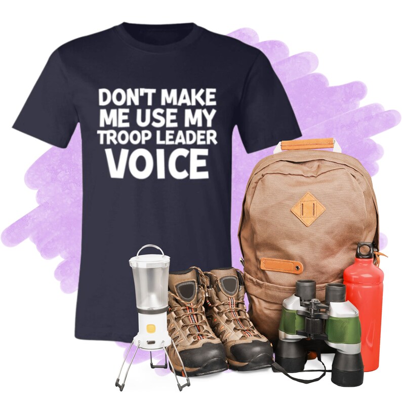 May include: A navy blue t-shirt with the text "Don't make me use my troop leader voice" in white. The t-shirt is next to a brown backpack, a pair of brown hiking boots, a green and black pair of binoculars, and a red water bottle.