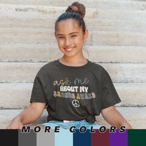 Peut inclure: Un t-shirt gris fonc&eacute; avec le texte "ask me about my bronze award" en lettres color&eacute;es. Un symbole de paix se trouve sous le texte. Le t-shirt est port&eacute; par une personne assise sur un escalier. L'image montre &eacute;galement un nuancier avec diff&eacute;rentes nuances de gris, de bleu, de vert et de violet.