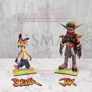 Puede incluir: Dos recortes de personajes del videojuego Jak y Daxter. Jak es una figura musculosa con pelo verde, gafas y armadura. Daxter es una criatura naranja con orejas grandes y vientre amarillo. Cada personaje está sobre una pequeña base con su nombre.