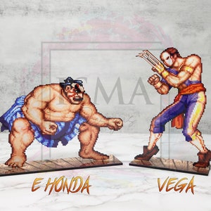 May include: Two pixel art figures from the Street Fighter video game series. E. Honda, a sumo wrestler, is in a fighting stance with his arms raised. Vega, a masked fighter, is in a fighting stance with his claws extended. Both figures are on wooden bases with their names printed below.