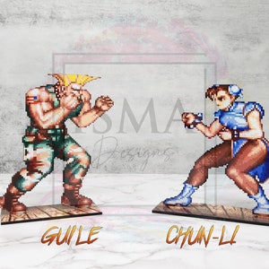 May include: Two pixel art figures from the Street Fighter video game series.  The figures are standing on wooden bases.  The figure on the left is Guile, a blonde male character wearing a green military uniform.  The figure on the right is Chun-Li, a female character with long black hair wearing a blue and white martial arts uniform.