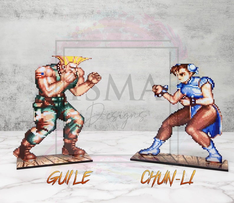 May include: Two pixel art figures from the Street Fighter video game series.  The figures are standing on wooden bases.  The figure on the left is Guile, a blonde male character wearing a green military uniform.  The figure on the right is Chun-Li, a female character with long black hair wearing a blue and white martial arts uniform.