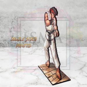 May include: A pixelated figure of a martial arts character in a white gi with a black belt, standing on a wooden platform. The figure is in a fighting stance with one hand raised to their chin. The figure is double-sided printed.