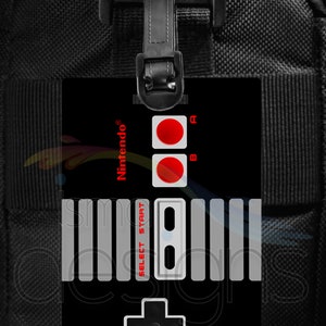 May include: Black luggage tag featuring a classic Nintendo controller design. The tag has red A and B buttons, a directional pad, and the words "SELECT START". The Nintendo logo is in red. The tag is attached to a black bag.
