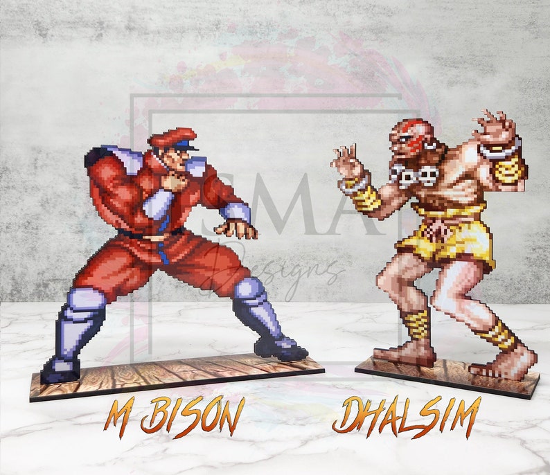 May include: Two pixel art figures from the Street Fighter video game series. The figures are standing on wooden bases. The figure on the left is M. Bison, a red and white character with a black hat. The figure on the right is Dhalsim, a yellow and brown character with a red headband.