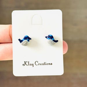May include: A pair of stud earrings shaped like small birds with dark blue heads, light blue accents, and textured grey wings. The earrings are attached to a white rectangular display card featuring the text Klay Creations in black font.