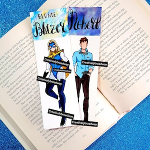 Puede incluir: Dos marcapáginas ilustrados sobre un libro abierto. Uno presenta a una superheroína en azul, amarillo y blanco. El otro muestra a un hombre con camisa azul y pantalón negro. Los marcapáginas tienen las palabras "Blonde Blazer" y "Robert" en escritura.