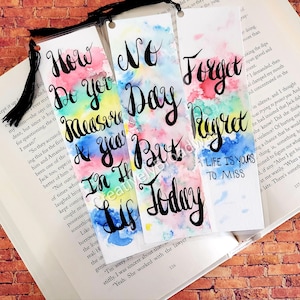 May include: Three watercolor bookmarks with inspirational quotes. The first bookmark is rainbow colored and says "How do you measure a year but in the life you live today". The second bookmark is blue and pink and says "No day but today". The third bookmark is green and blue and says "Forget regret, life is yours to miss".