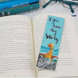 Könnte beinhalten: Ein blaues und weißes Lesezeichen mit dem Text "If You Knew My Story" in schwarzer Kursivschrift. Das Lesezeichen zeigt eine Aquarellmalerei einer Frau in einem gelben Kleid und einem Strohhut, die auf einer felsigen Klippe steht und auf das Meer blickt.