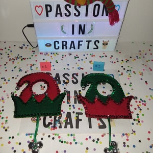 May include: Two felt elf hats with jingle bells, one red and green, the other green and red, with the words "Passion in Crafts" on a light box behind them.