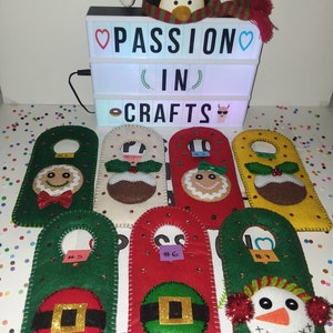 May include: Seven felt Christmas door hangers with different designs. The designs include a gingerbread man, a Christmas pudding, a snowman, and Santa Claus. The hangers are decorated with glitter and sequins. The hangers are numbered 1 through 7. The sign above the hangers reads "PASSION IN CRAFTS".
