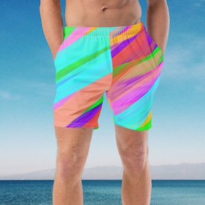 May include: A pair of colorful swim trunks with a bright blue, pink, green, and yellow abstract pattern. The trunks are on a man standing on a sandy beach with a blue boat in the background.