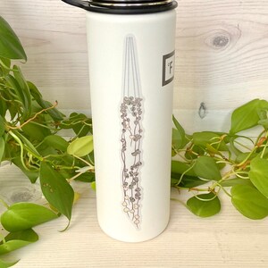 May include: White stainless steel water bottle with a black lid and a black carrying handle. The bottle has a graphic of a hanging plant with green leaves and white flowers.