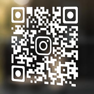 May include: White QR code sticker featuring an Instagram logo at its centre. The code is made up of black and white squares, displayed on a glass surface. The background is blurred, implying an outdoor setting.