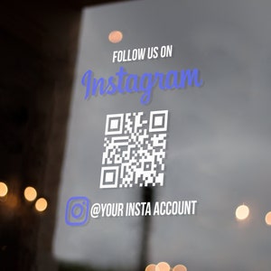 May include: A white sticker on a glass door with the text "FOLLOW US ON Instagram" in blue text. A black and white QR code is below the text. Below the QR code is a white Instagram icon with the text "@YOUR INSTA ACCOUNT" in white text.