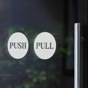 May include: Two white round signs with black text on a glass door. The sign on the left says "PUSH" and the sign on the right says "PULL".