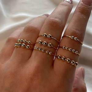 Könnte beinhalten: Ein Satz von fünf goldenen Perlenringen, die auf einem Finger übereinander liegen. Die Ringe bestehen aus dünnem Metall mit kleinen, gleichmäßig verteilten goldenen Perlen.