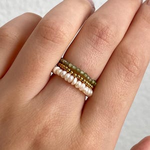 Könnte beinhalten: Nahaufnahme von drei Perlenringen an einer Hand. Ein Ring hat kleine weiße Perlen, ein anderer grüne Perlen und der dritte ist goldfarben. Die Ringe sind gestapelt, wodurch ein geschichteter Look entsteht. Der Hintergrund ist eine schlichte weiße Oberfläche.
