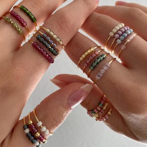 Può includere: Una collezione di delicati anelli d'oro, ognuno ornato da una fila di perline colorate. Gli anelli presentano una varietà di colori di perline, tra cui verde, rosso, bianco e viola. Gli anelli sono mostrati su più dita.