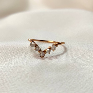 Könnte beinhalten: Ein zarter, roségoldener Ring mit V-förmigem Design. Der Ring hat fünf kleine, hellfarbige Edelsteine, die entlang des V angeordnet sind. Der Ring wird auf einem weichen, weißen Stoffhintergrund präsentiert.