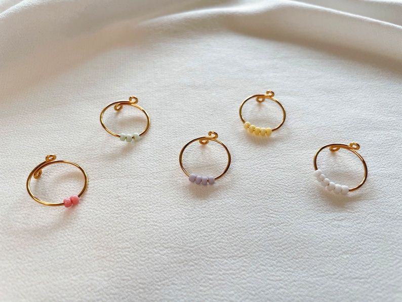 May include: Five gold rings with different colored beads. The rings are all the same size and shape. The beads are in a variety of colors, including pink, white, light green, and light purple.