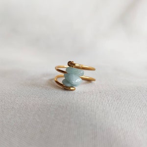 Puede incluir: Anillo de oro con tres piedras de color azul claro apiladas. El anillo está hecho de una banda delgada y retorcida.