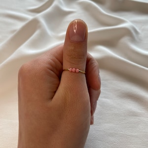 May include: A gold ring with a row of small pink beads. The ring is worn on a person's thumb.