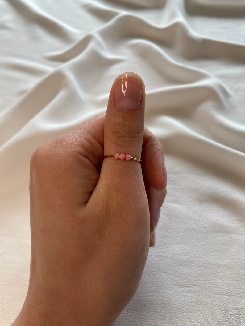 May include: A gold ring with a row of small pink beads. The ring is worn on a person's thumb.
