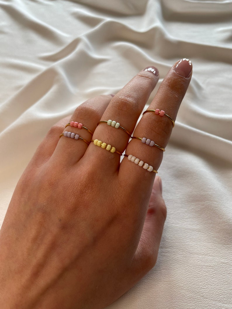May include: Set of five gold rings with colorful beads. The rings are stacked on a finger. The beads are in shades of pink, yellow, and white.