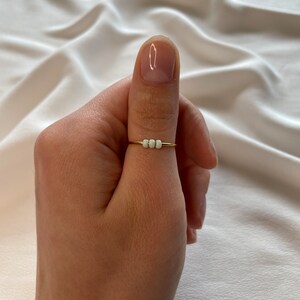 May include: A gold ring with three white beads on a person's thumb.